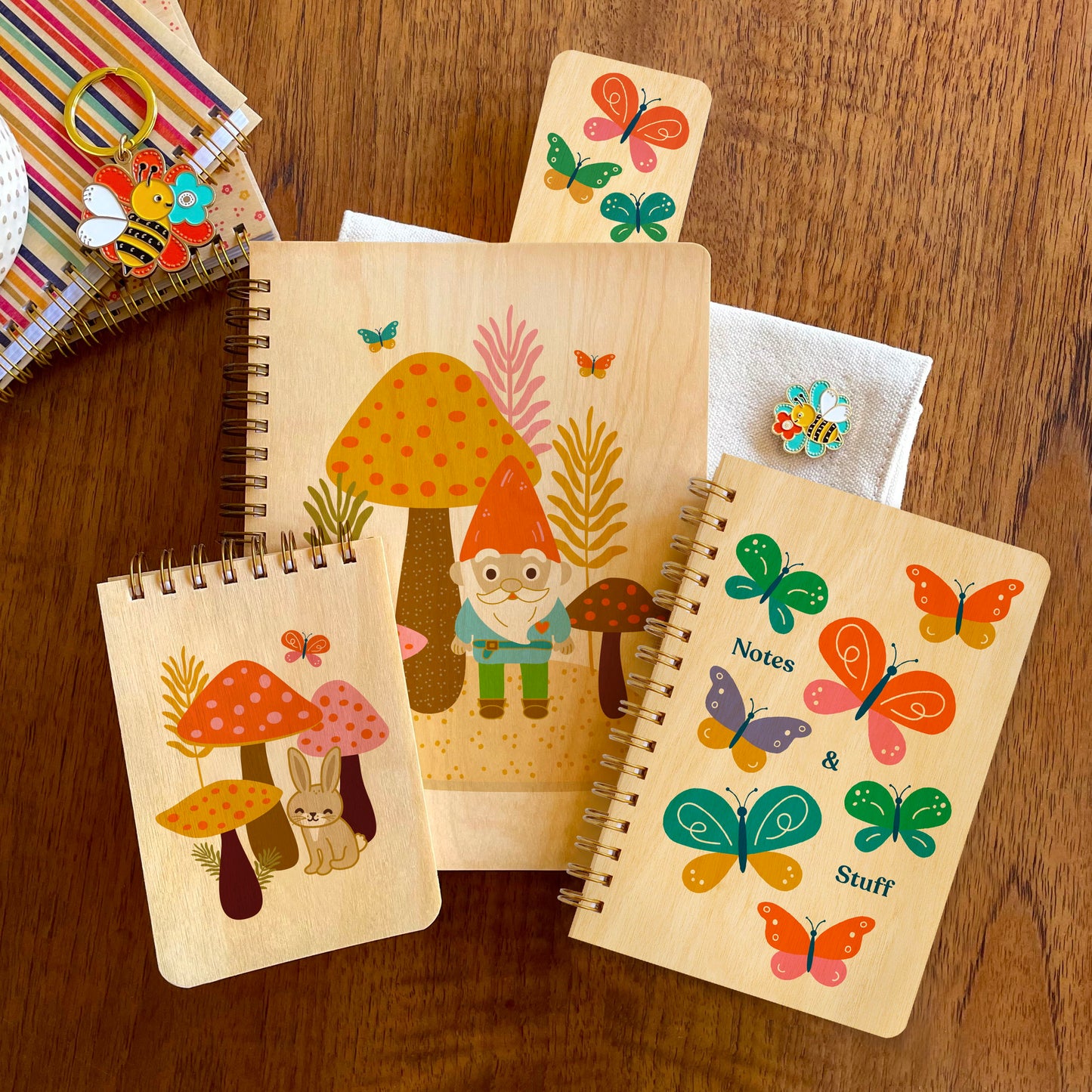 Collection of nature-themed wood notebooks, including a woodland gnome, colorful butterflies and rabbit with mushrooms. Also shown are a butterflies bookmark and a bumble bee enamel pin.