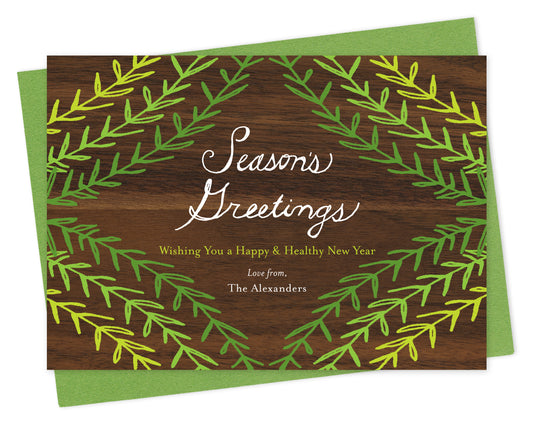 Customizable paper holiday card with green sprig design, "Season's Greetings" sentiment and a personal message on a printed walnut wood grain background. Shown with a green envelope.