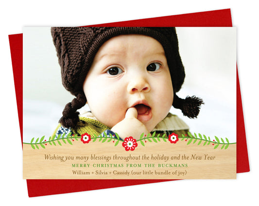 Customizable paper holiday card featuring a photo with a floral garland and printed birch wood grain border at the bottom and a personal greeting. Shown with a red envelope.