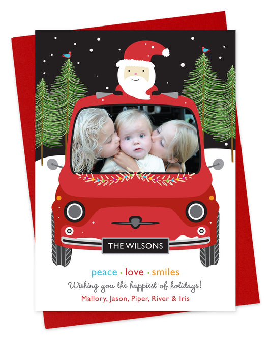 Customizable paper holiday card featuring your photo in the windshield of a red car with santa riding on top. Features family name on the license plate and a personal greeting at the bottom of the card. Shown on a red envelope.