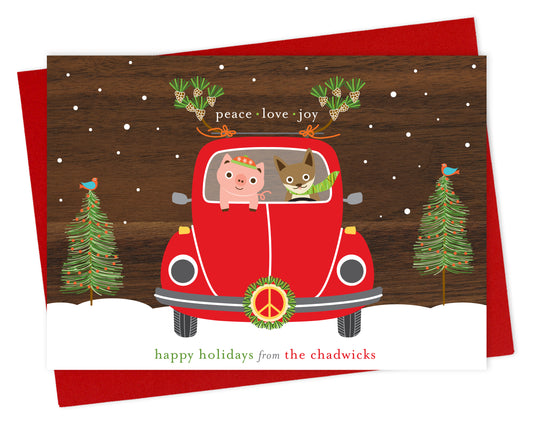 Customizable paper holiday card with fun illustration of a pig and fox riding in a red car with peace symbol and wreath on the front of the car with a printed walnut wood grain sky. Card features “Peace Love Joy” sentiment and a personal greeting. Shown with a red envelope.