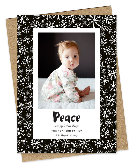 Customizable paper holiday card featuring a family photo with “Peace” text and a personal greeting near the bottom, framed by a black border with a snowflake pattern. Shown with a kraft envelope.