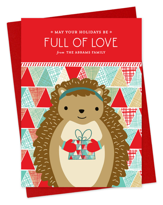 Customizable paper holiday card featuring a cute gift-giving hedgehog, “May Your Holidays Be Full Of Love” text and a personal greeting on a colorful, patchwork background. Shown with a red envelope.