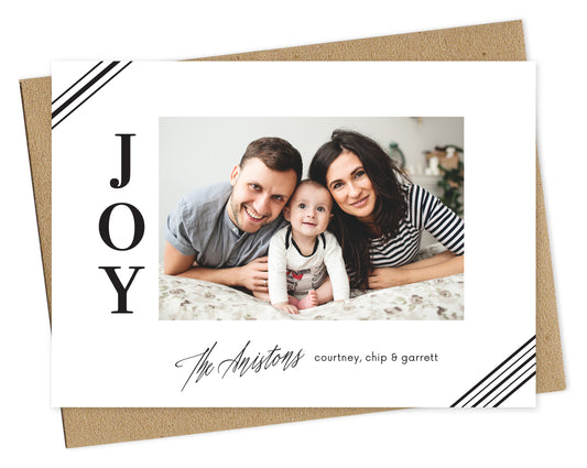 Customizable paper holiday card with a photo framed by white, corner stripes, the word “JOY” on the left and a personal greeting on the bottom, all printed in black ink. Shown with a kraft envelope.