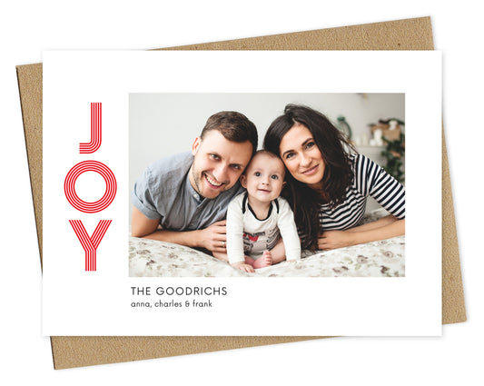 Customizable paper holiday card featuring your photo bordered by white with the word "JOY" in red on the left and a personal greeting in black on the bottom. Pictured with a kraft envelope.