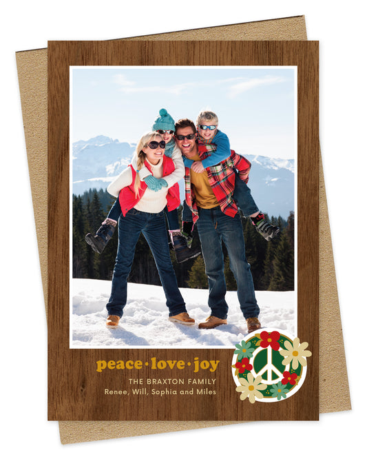 Customizable paper holiday card featuring a family photo with a printed walnut wood grain border and “peace love joy” text and a peace sign surrounded by flowers near the bottom. Shown with a kraft envelope.