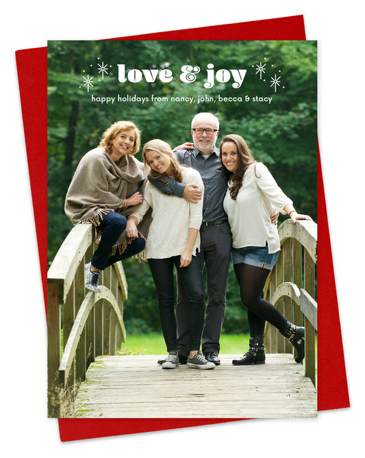 Customizable paper holiday card featuring a full photo with "love & joy” text and sparkle illustrations near the top, shown with a red envelope.