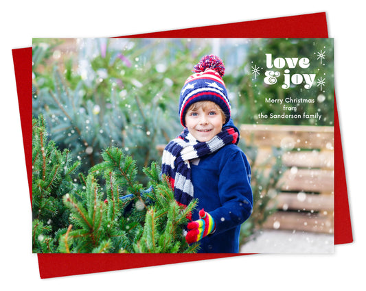 Customizable paper holiday card featuring a full landscape photo with "love & joy” text and sparkle illustrations near the top, shown with a red envelope.