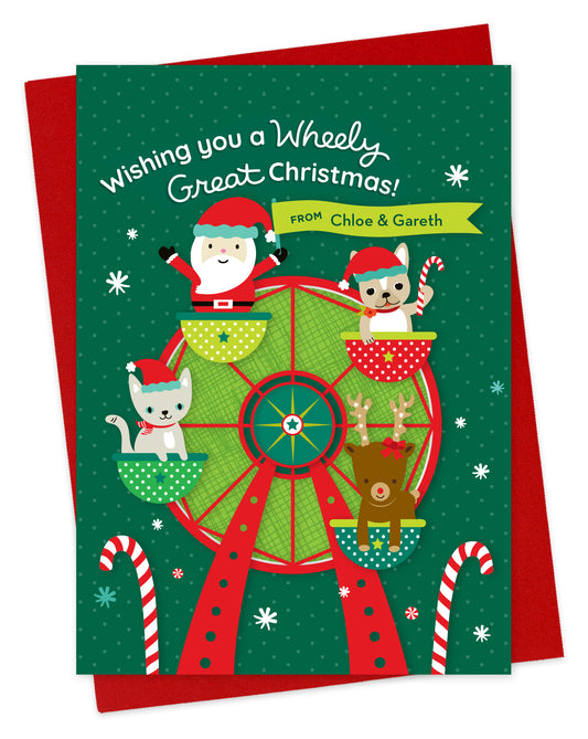 Customizable paper holiday card with sentiment "Wishing you a Wheely Great Christmas!” Features a festive holiday ferris wheel with a cute Santa waving a banner that contains an area for a personal greeting. Ferris wheel also has a kitty cat wearing a Santa hat and scarf, a french bulldog wearing a Santa hat holding a candy cane and a reindeer with a bow. Shown with a red envelope.