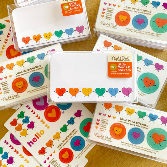 Happy Hearts Little Cards & Stickers boxed sets featuring mini note cards decorated with a row of smiling rainbow hearts and coordinating vinyl stickers with colorful hearts and cheerful designs. The clear boxes are arranged on a wooden surface.
