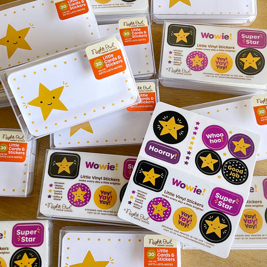 Overhead product photo of Night Owl Paper Goods Gold Star Little Cards & Stickers boxed sets, featuring smiling gold star mini note cards and coordinating colorful vinyl stickers with encouraging phrases like “Wowie!,” “Hooray!,” “Super Star” and “Good Job.” Gift-ready clear boxes are arranged on a wooden surface.