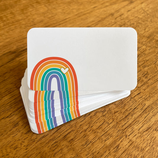 A stack of small white rainbow note cards with rounded corners, featuring a colorful rainbow with an overlapping white heart.