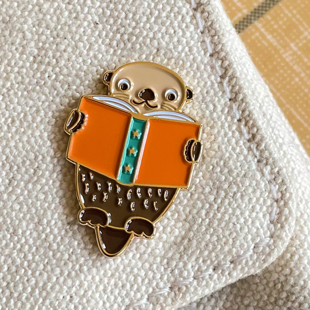 Enamel pin of an otter reading a book on a canvas background.