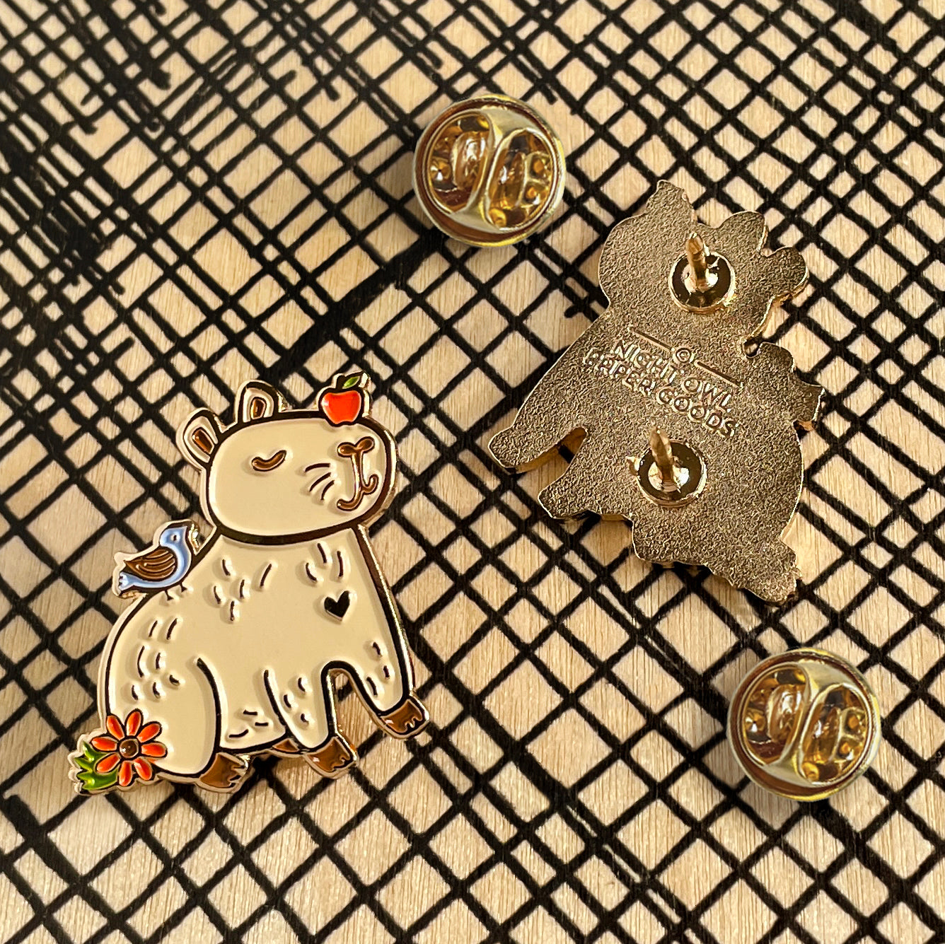 Views of front and back of gold metal Capybara enamel pin, alongside two butterfly clasps. Front of pin features a smiling capybara with an apple on its head, as well as bird and flower details. Back of pin shows dual posts and Night Owl Paper Goods logo.