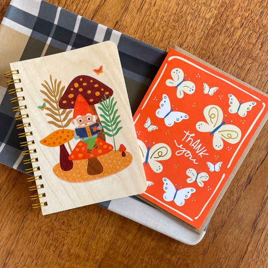 Wood notebook with happy gnome sitting on a mushroom while reading a book in a forest setting. A red thank you card features butterflies and shiny gold accents.