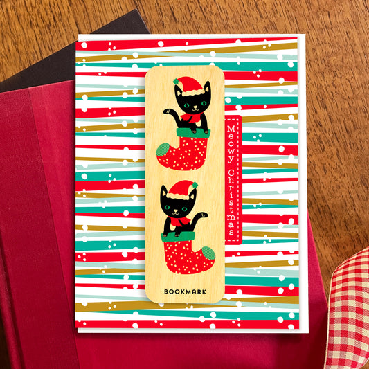 Folded Christmas greeting card with colorful red, green and gold striped background, “Meowy Christmas” text and a removable birch wood bookmark featuring two black cats in red Christmas stockings. Pictured with a white envelope and sitting atop a book on a wooden table with a red gingham ribbon.