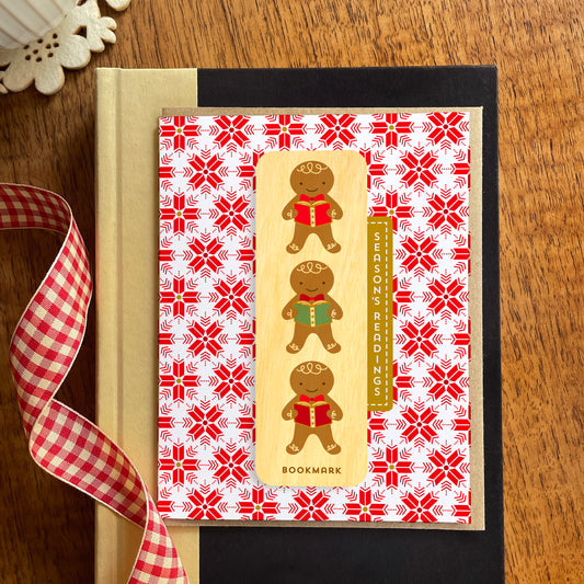 Folded holiday greeting card with red and white snowflake pattern, “Season’s Readings” text and a removable birch wood bookmark featuring three gingerbread cookies holding books. Pictured with a kraft envelope and sitting atop a book on a wooden table with a red gingham ribbon.