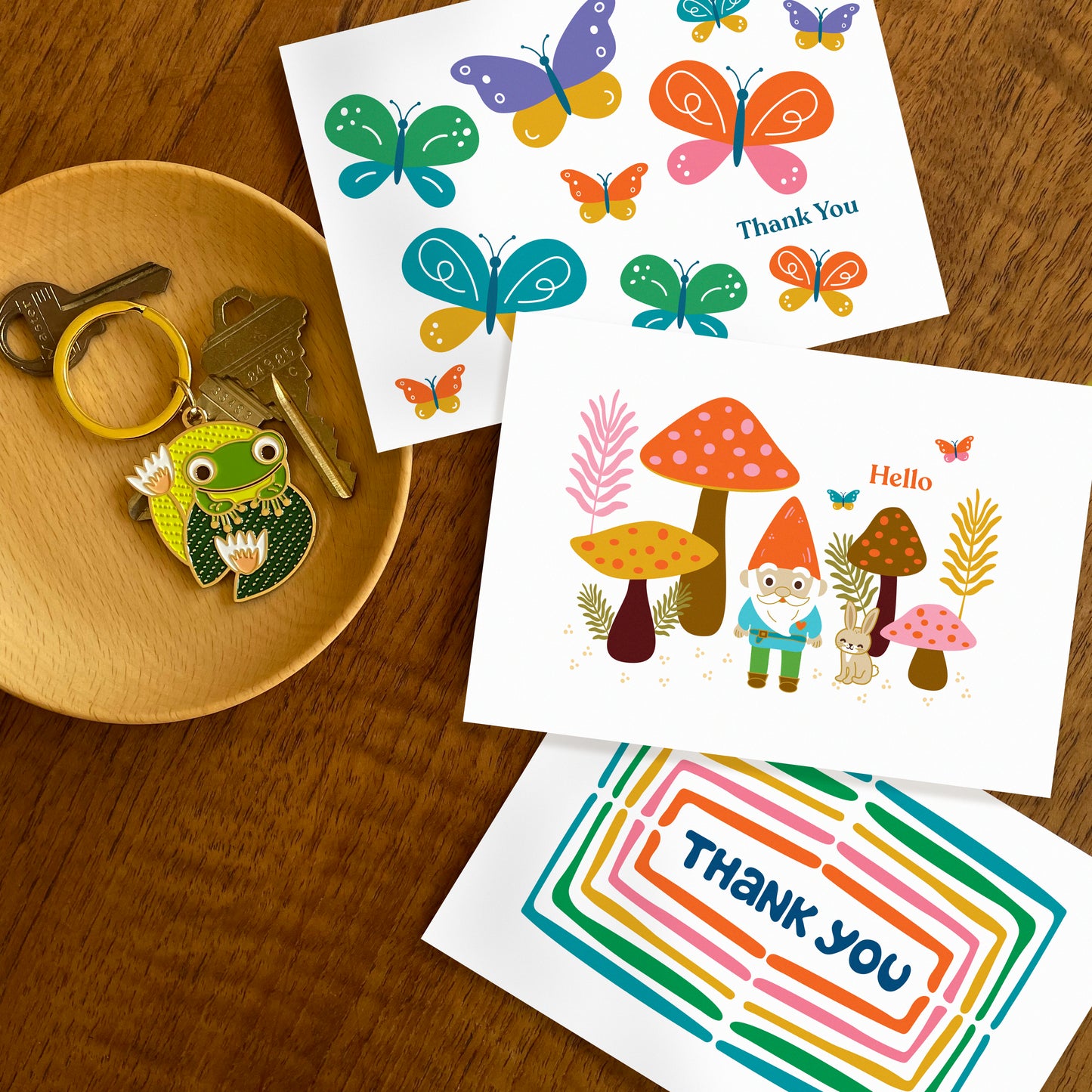 Three folded thank you cards colorful butterflies, gnome in forest with mushrooms and rabbit, and a rainbow of colors surrounding a hand drawn word 'thank you'. Frog on lily pad keychain also featured