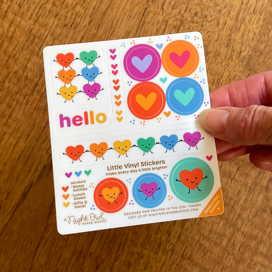Hand holding a sheet of colorful heart and rainbow-themed vinyl stickers on a white background. The stickers include hearts and smiling hearts in red, orange, yellow, green, blue and purple, a word sticker that says "hello" in multicolor letters. Text on the sheet reads "Little Vinyl Stickers - make every day a little brighter - Decorate water bottles, lunch boxes, gifts & more - Night Owl Paper Goods - Designed and printed in the USA"