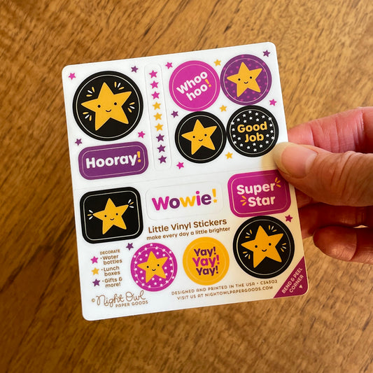Hand holding a sheet of cute star-themed kiss-cut vinyl stickers on a white background. The stickers feature smiling golden stars and colorful designs with encouraging sayings like “Hooray!”, “Whoo hoo!”, “Good Job,” “Wowwie!”, “Super Star” and “Yay! Yay! Yay!” in gold, purple, pink and black. Text on the sheet reads “Little Vinyl Stickers - make every day a little brighter - Decorate water bottles, lunch boxes, gifts & more - Night Owl Paper Goods - Designed and printed in the USA.”