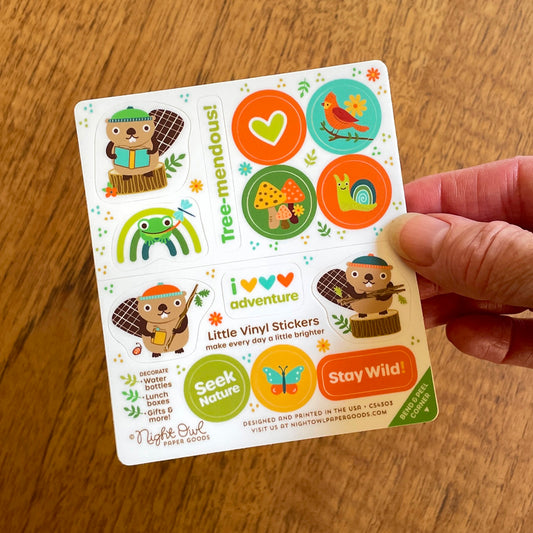 Hand holding a sheet of cute woodland animal kiss-cut vinyl stickers on a white background. The stickers feature beavers, a frog, a heart, mushrooms, a bird, a snail, a butterfly and sayings like "Tree-mendous," "Seek Nature," and "Stay Wild!" in warm earthy colors like orange, green, brown and gold. Text on the sheet reads "Little Vinyl Stickers - make every day a little brighter - Decorate water bottles, lunch boxes, gifts & more - Night Owl Paper Goods - Designed and printed in the USA"