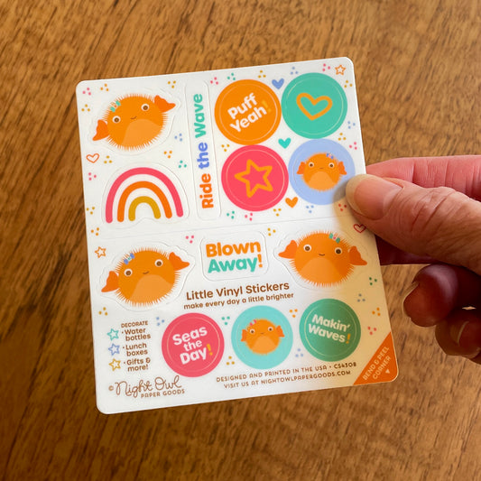 Hand holding a sheet of cute ocean-themed kiss-cut vinyl stickers on a white background. The stickers feature orange puffer fish, a rainbow, hearts, stars and colorful sayings like "Puff Yeah," "Blown Away!," "Seas the Day!" and "Makin' Waves!" in bright orange, pink, teal, blue and yellow. Text on the sheet reads "Little Vinyl Stickers - make every day a little brighter - Decorate water bottles, lunch boxes, gifts & more - Night Owl Paper Goods - Designed and printed in the USA."