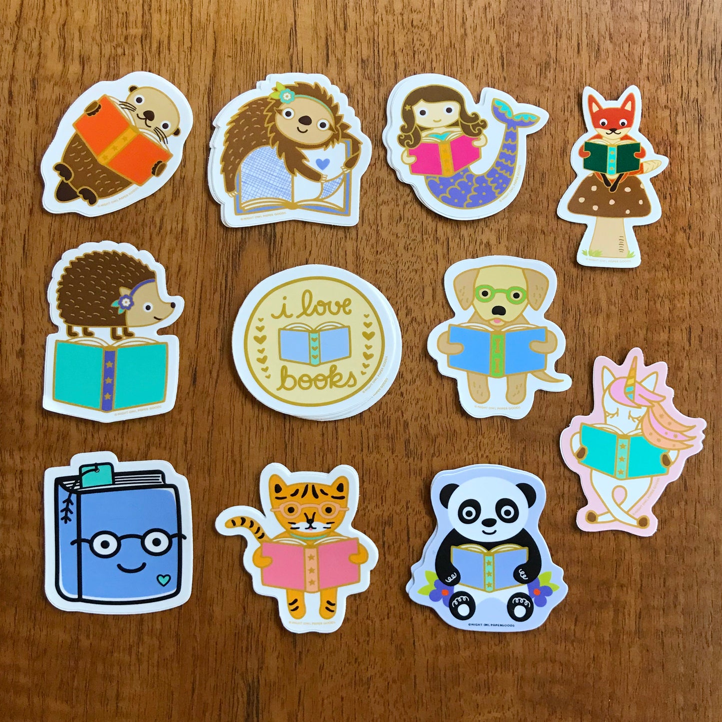 11 book lover stickers, including a reading golden retriever, a reading sloth, a reading unicorn, a reading otter, a hedgehog standing on a book, an otter holding a book, a reading tiger, a mermaid holding a book, a reading panda bear, a happy book wearing glasses and a round sticker that features an open book and "I love books" text.