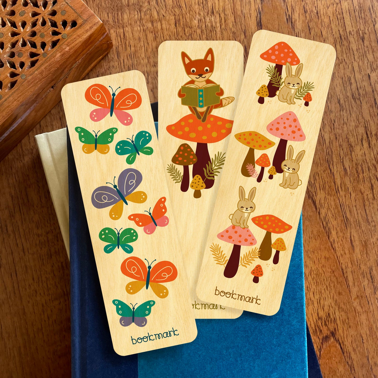 Group of three bookmarks on a stack of books. Bookmarks feature colorful butterflies, a fox sitting on a mushroom reading a book and bunny rabbits with mushrooms