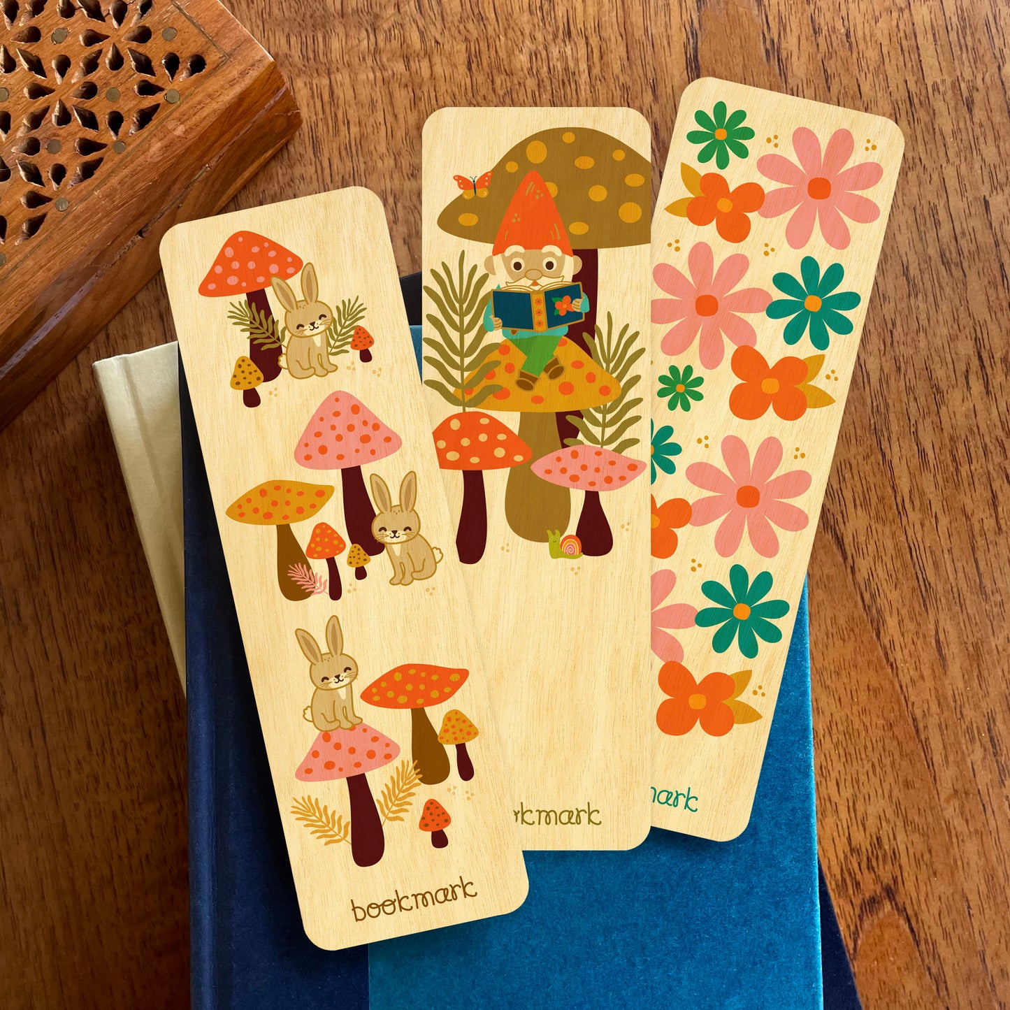 Group of three wooden bookmarks sitting on a stack of books. Bookmarks feature rabbits, mushrooms, a gnome reading and a colorful flower pattern