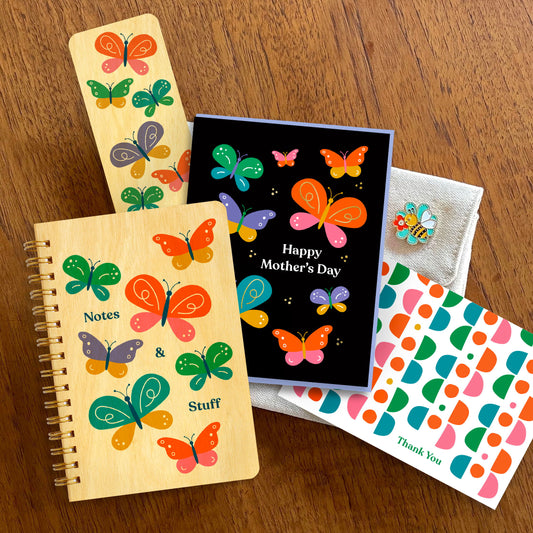 Wood notebook and bookmark with colorful butterflies, black "Happy Mother's Day" greeting card with colorful butterflies, bumble bee enamel pin and white thank you card with colorful pattern.