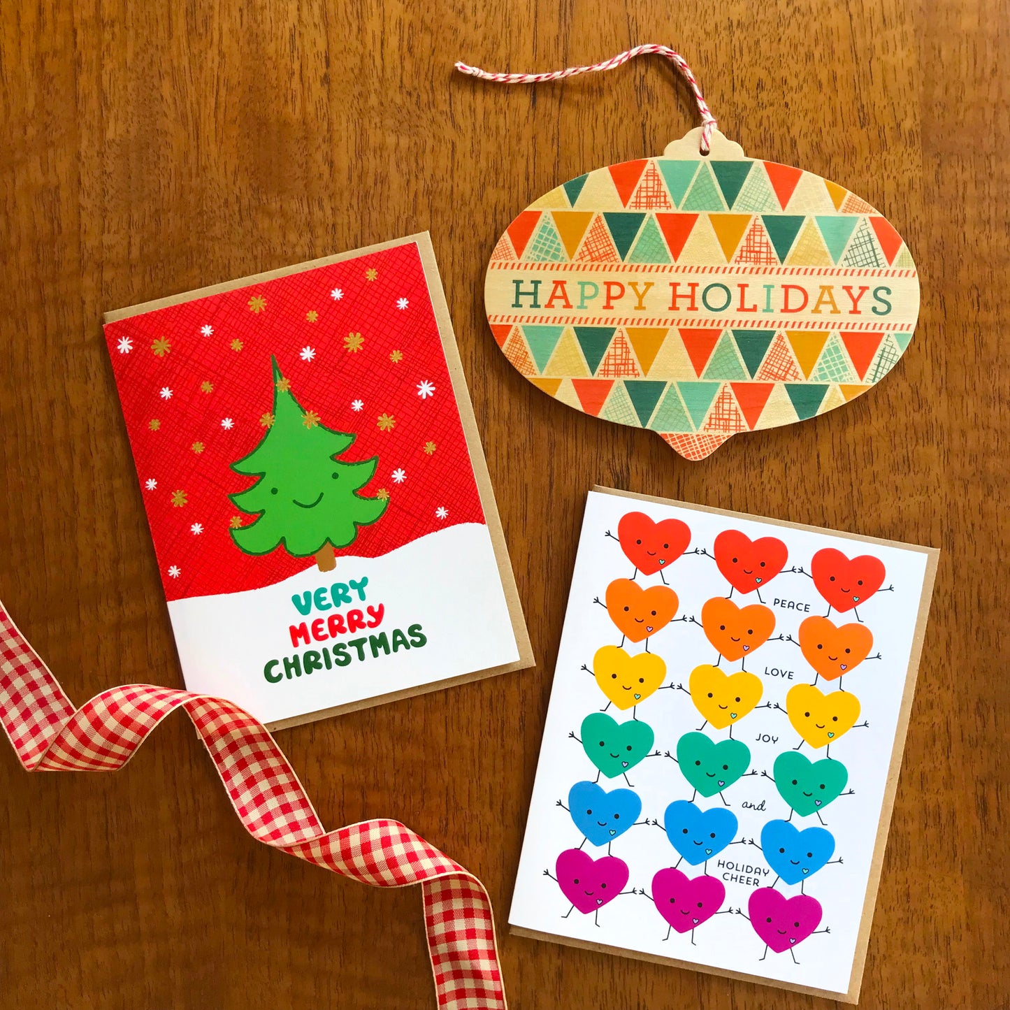 Group of three holiday cards featuring a smiling green tree with text that reads 'Very Merry Christmas', a wood ornament card shaped like a wide bulb text reads ' happy holidays' and rainbow happy hearts dancing with text reads 'peace love joy and holiday cheer'