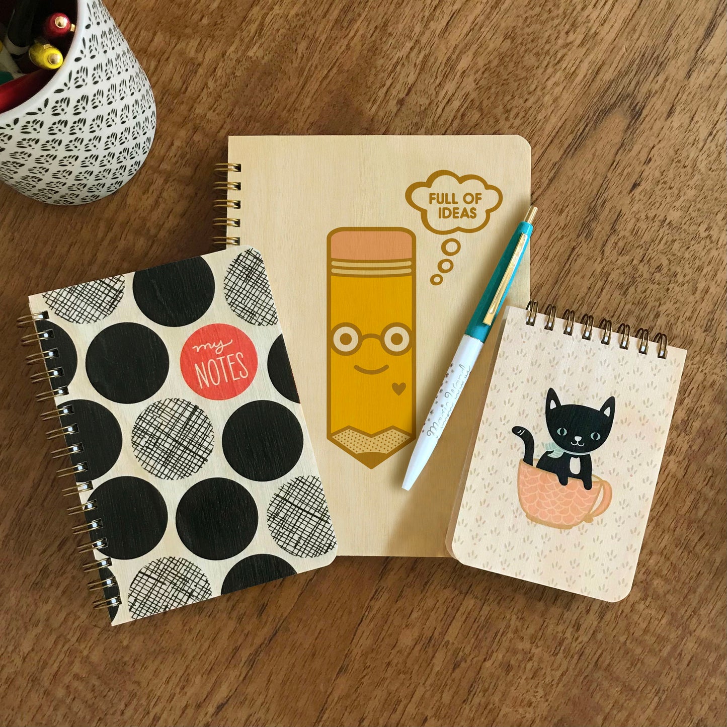 Big Dots Notes Wood Notebook, Full of Ideas Wood Journal and Cuppa Kitty Wood Mini Notepad side-by-side on a wood table. A white and aqua ballpoint click pen sits atop the journal.