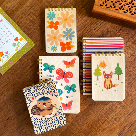 Group of notebooks and mini notepads with wood covers featuring colorful flowers, lines, butterflies, a beaver and a fox.