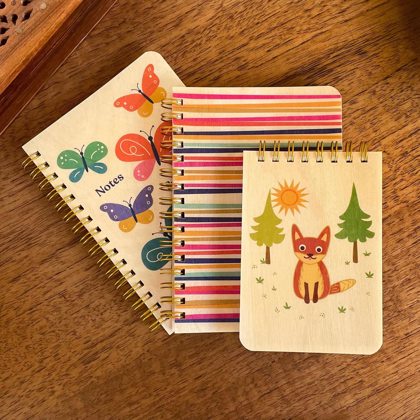 Three small notebooks with wood covers and different colorful designs on top of a desk