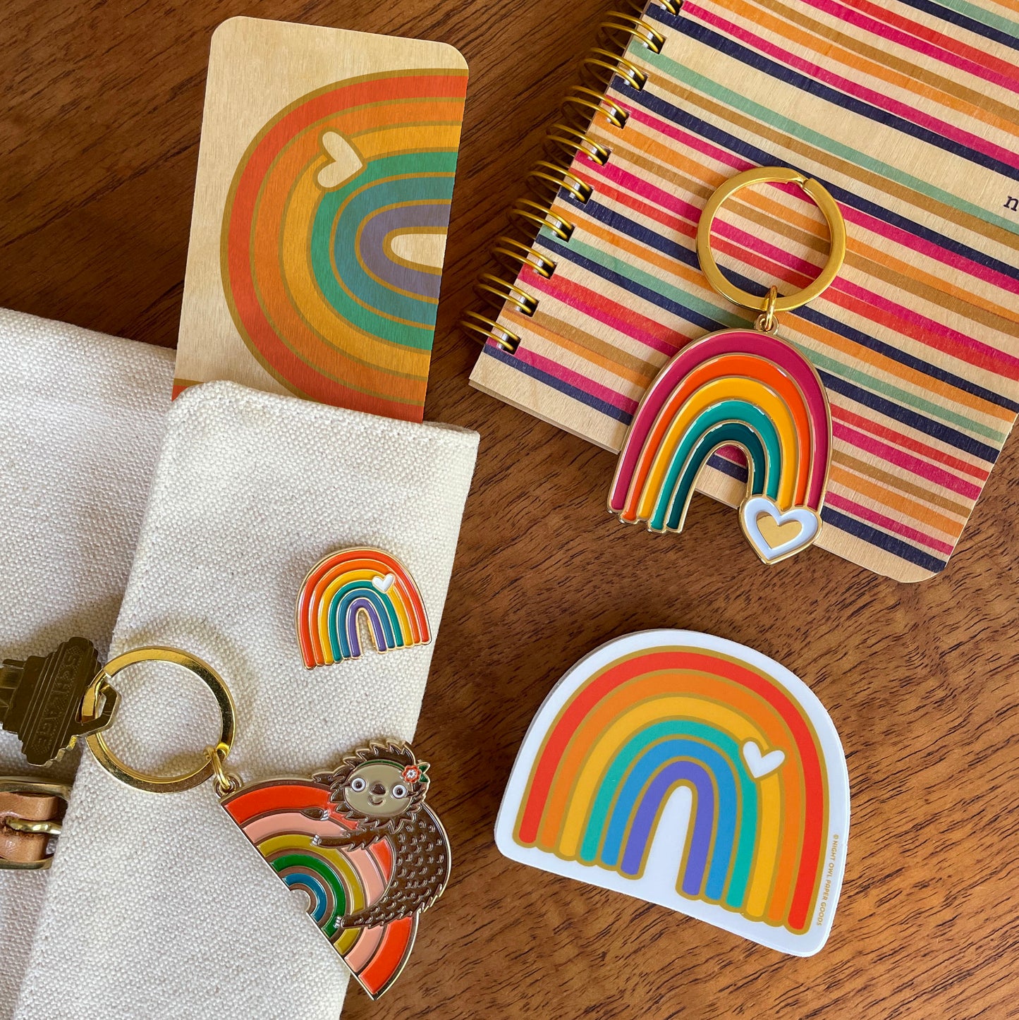 Group photo with a colorful assortment of rainbow products. Wooden bookmark, striped notebook, keychains, enamel pin and sticker