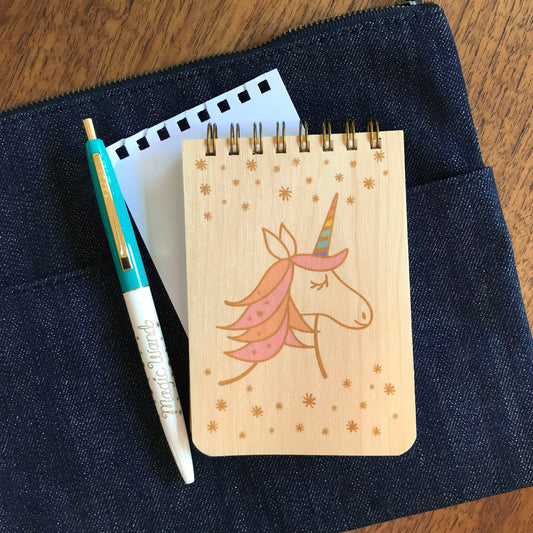 Spiral-bound wood mini notepad with birch wood covers featuring an enchanting unicorn with a flowing peach and pink mane and pastel rainbow-striped horn surrounded by scattered stars. Next to it sits a ballpoint click pen and a torn out sheet of blank white inner paper.