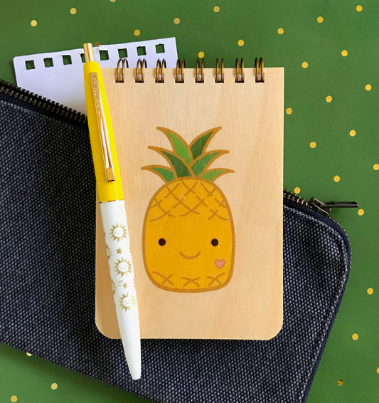 Spiral-bound wood mini notepad with birch wood covers featuring a smiling kawaii-style pineapple character with a tiny pink heart and green leafy top. Next to it sits a ballpoint click pen with suns and a torn out sheet of blank white inner paper.
