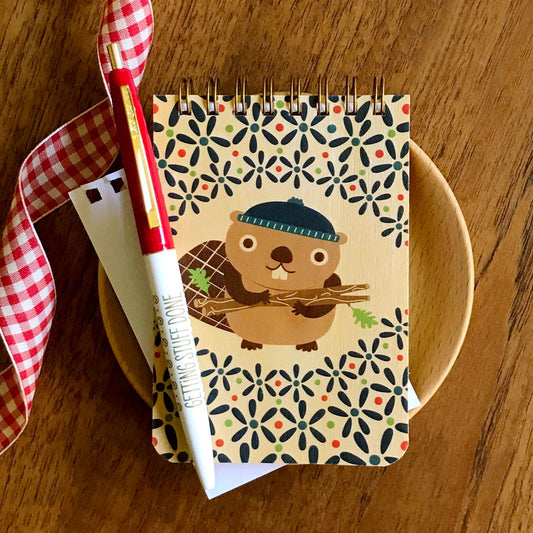 Spiral-bound wood mini notepad with birch wood covers featuring a smiling beaver in a navy blue beanie carrying branches surrounded by a bold navy, red and green floral pattern. Under the notepad is a torn-out sheet of white inner paper and on top of the notepad sits a red and white Getting Stuff Done ballpoint pen.
