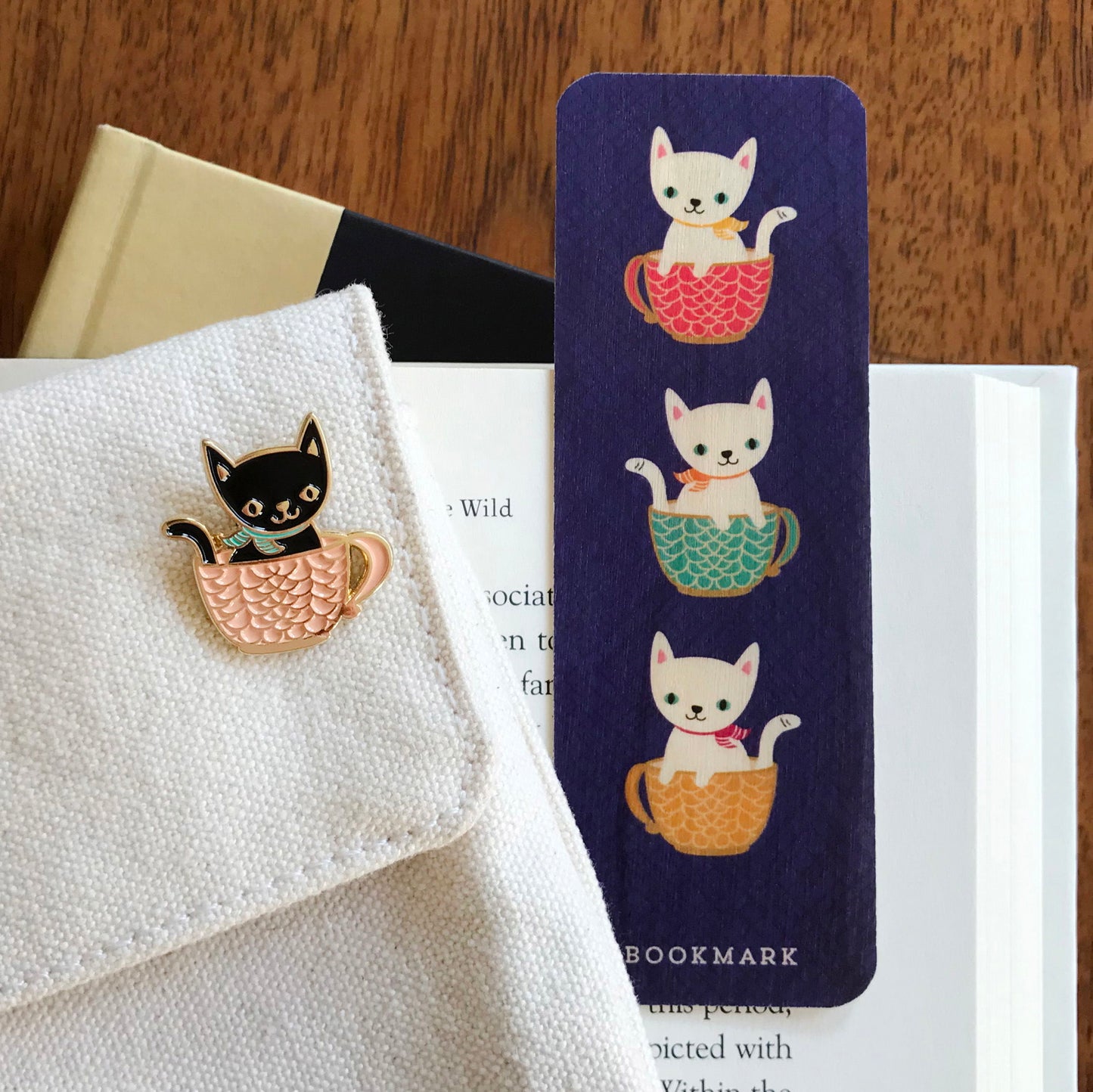 Enamel pin of a black cat in soft pink coffee mug and a blue wood bookmark with 3 white cats in colorful coffee cups, sitting on an open book