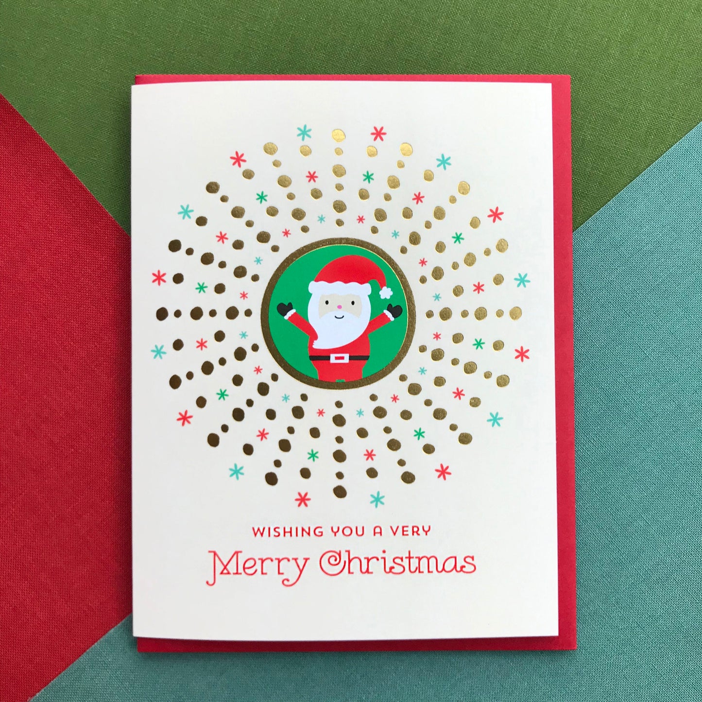 Photo of Christmas card sitting on colorful background, greeting card has Santa Claus illustration, gold foil details and festive text on a soft white background