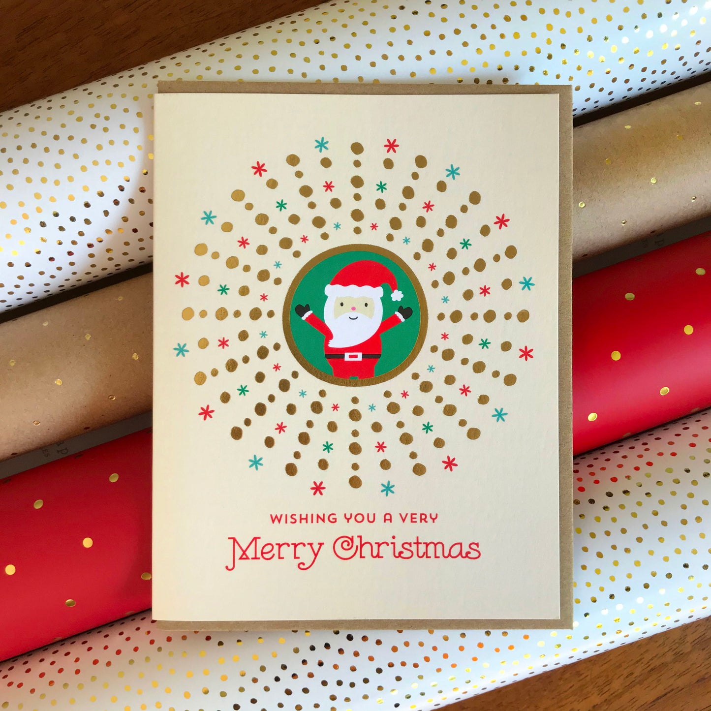 Santa Gold Foil Holiday Card featuring a cheerful Santa Claus in a green circle surrounded by a gold foil dot sunburst and red and green snowflakes on a cream background with the greeting "Wishing You a Very Merry Christmas." Shown with a kraft envelope atop rolls of festive wrapping paper.