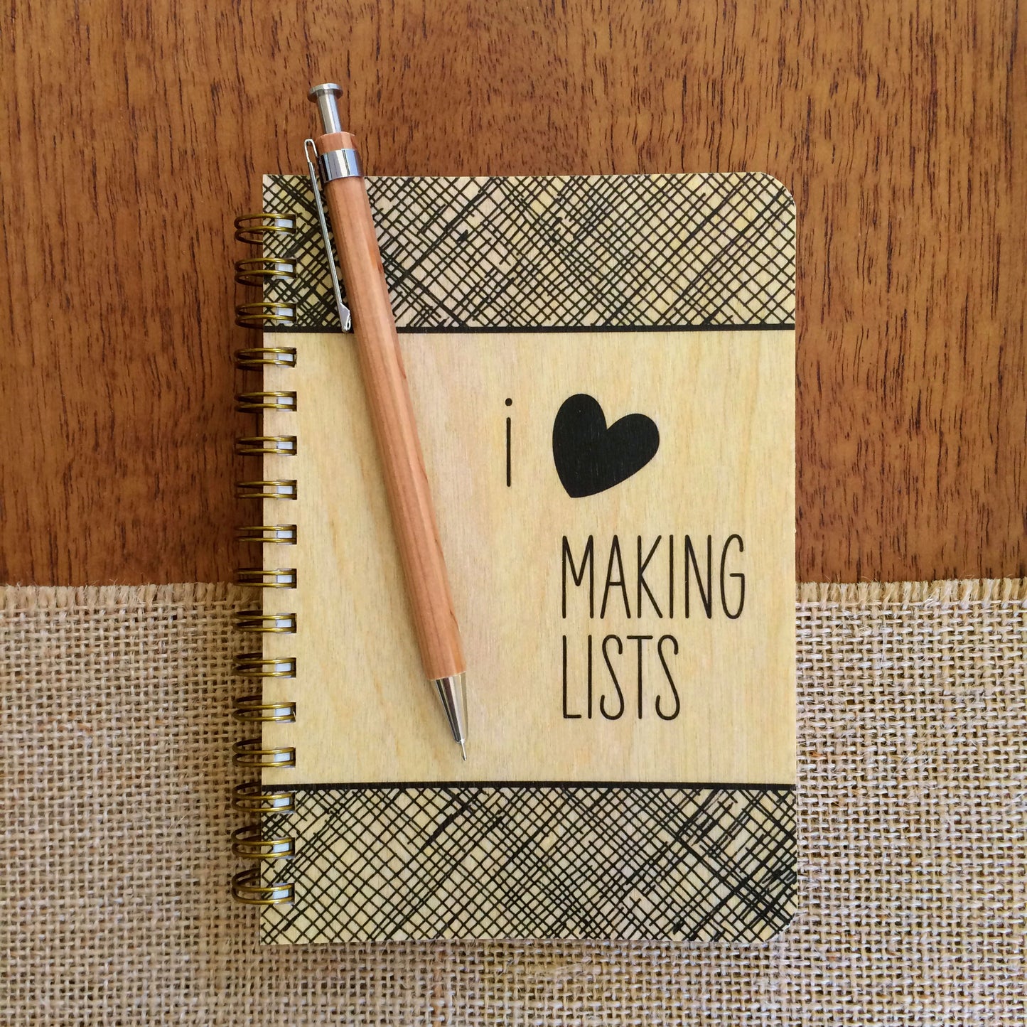 Spiral-bound wood notebook with birch wood covers featuring "I ♥ Making Lists" text and a black crosshatch border sitting on a wood table. A pencil sits atop the notebook.