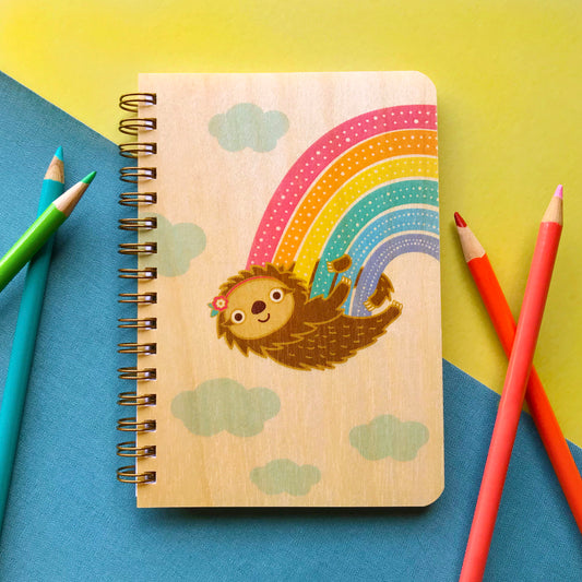 Spiral-bound wood notebook with birch wood covers featuring a sloth with a floral headband hanging from a rainbow among clouds. Notebook sits among colored paper and pencils.