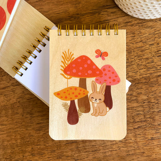 Spiral-bound wood mini notepad with birch wood covers featuring a happy bunny nestled among spotted mushrooms with a butterfly and wildflowers sits upon an open mini notepad that shows blank white inner pages.