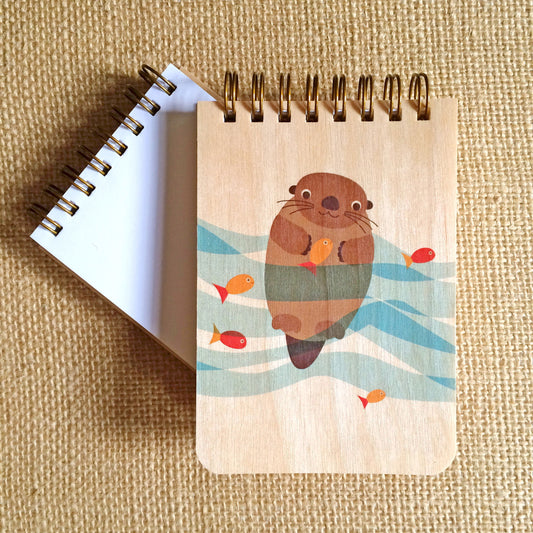 Spiral-bound wood mini notepad with birch wood covers featuring a happy otter floating in teal wavy water with orange fish swimming nearby sits upon an open mini notepad that shows blank white inner pages.