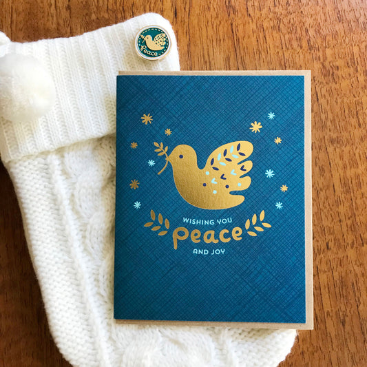White holiday stocking with blue and gold dove enamel pin text reads 'peace' and a blue holiday greeting card with a gold foil dove and text 'Wishing you peace and joy'