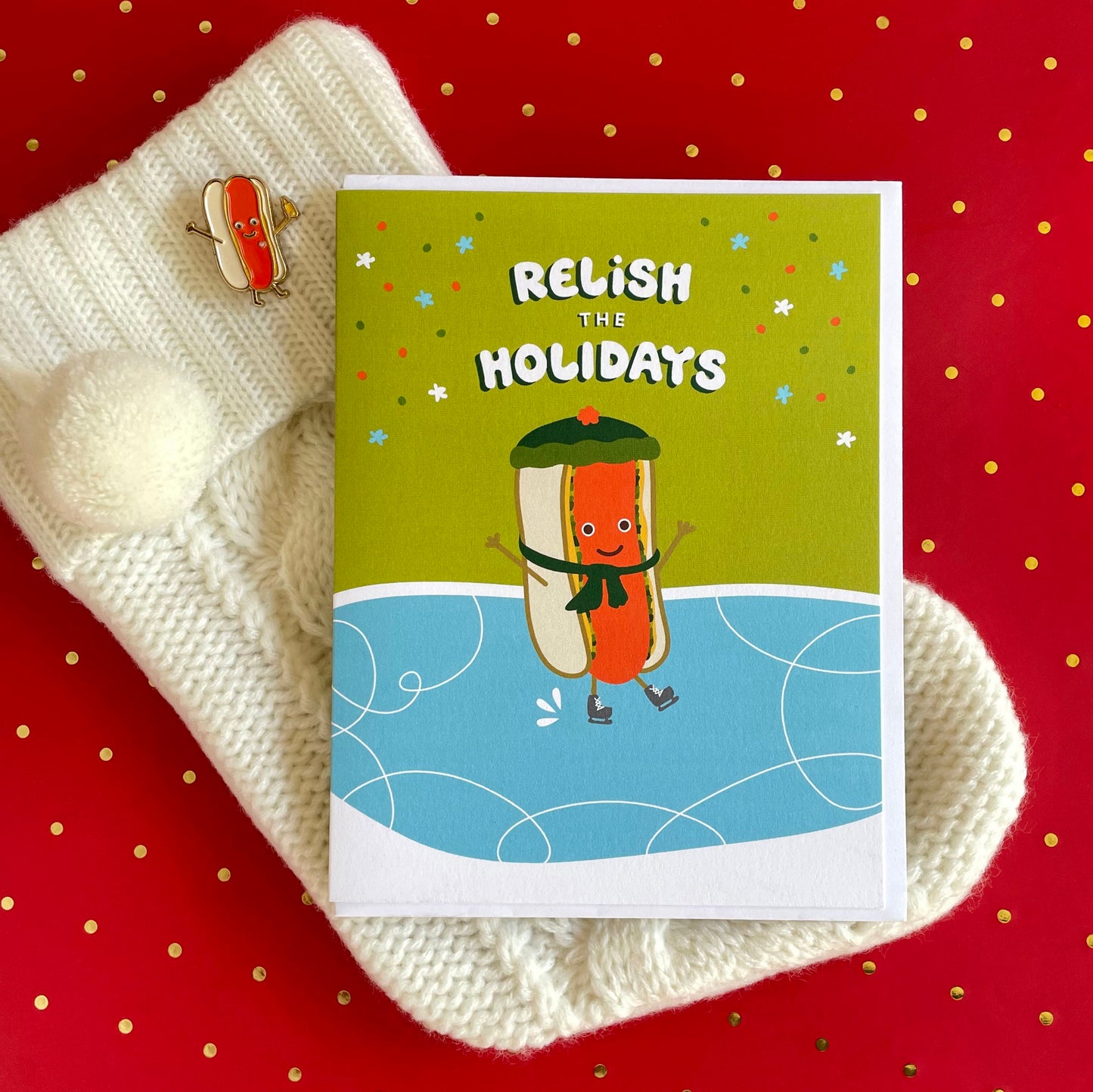 Hot Dog Holiday Card featuring a smiling hot dog in a green scarf and ice skates skating on a blue ice rink against an olive green background with a "Relish the Holidays" greeting. Shown with a white envelope alongside a Hot Dog enamel pin atop a Christmas stocking and a festive red background.