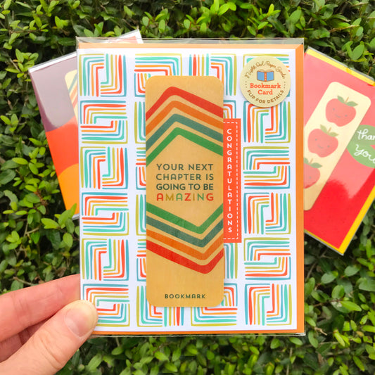 Congrats Chapter Bookmark Congratulations Card shown packaged in a clear sleeve with a descriptive sticker that reads Night Owl Paper Goods Bookmark Card Flip for Details. Two additional cards partially appear in the background against a leafy backdrop.