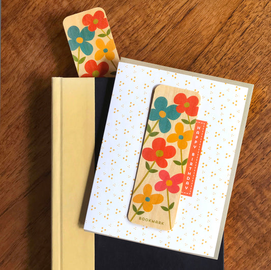 Folded birthday greeting card with a white background and golden confetti dot pattern, Happy Birthday text and a removable birch wood bookmark featuring bold retro flowers in teal, orange, pink and gold. Pictured with a kraft envelope and shown alongside a book with another floral bookmark sticking out the top.