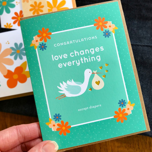 Baby shower card with stork and text 'Congratulations love changes everything except diapers' held by a hand.